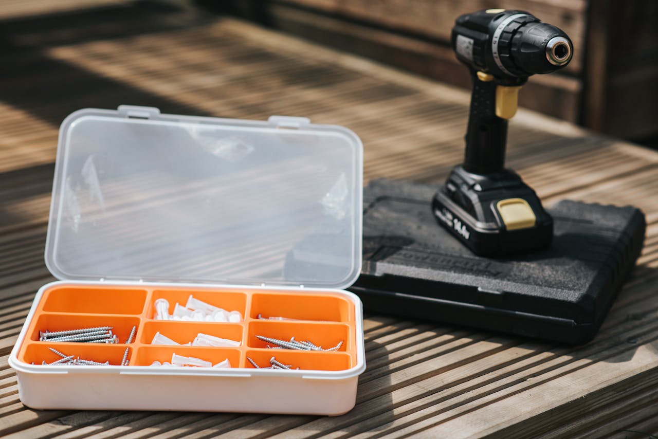 Cordless electric screwdriver placed on case on wooden surface near screw set with bolts and nails on street on blurred background